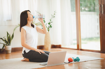 Healthy young Asian woman drinking from her water bottle while using laptop practicing yoga at home