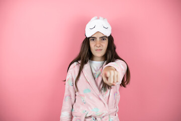 Pretty girl wearing pajamas and sleep mask over pink background pointing with finger something on the front