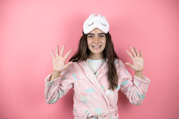 Obraz na płótnie Canvas Pretty girl wearing pajamas and sleep mask over pink background showing and pointing up with fingers number ten while smiling confident and happy