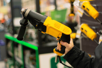 Man holds a yellow drill in his hands for repair work on the background of showcases in a hardware store.