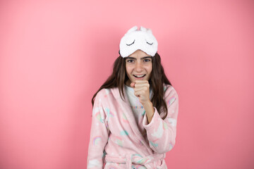 Pretty girl wearing pajamas and sleep mask over pink background with her hand to her mouth coughing