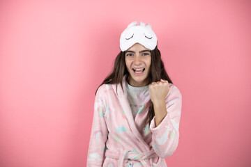 Pretty girl wearing pajamas and sleep mask over pink background angry and mad raising fist frustrated and furious while shouting with anger.
