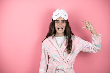 Pretty girl wearing pajamas and sleep mask over pink background smiling and confident gesturing with hand doing small size sign with fingers looking and the camera. Measure concept.