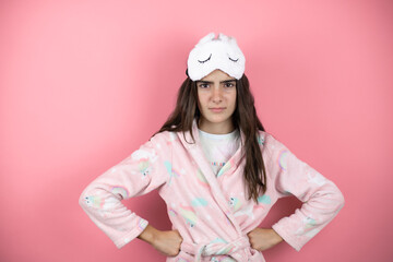 Pretty girl wearing pajamas and sleep mask over pink background skeptic and nervous, disapproving expression on face with arms in waist