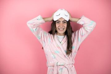 Pretty girl wearing pajamas and sleep mask over pink background suffering from headache desperate and stressed because pain and migraine with her hands on head