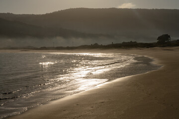 beautiful curved golden sandy beach with glistening waves in the morning light and fog in the...
