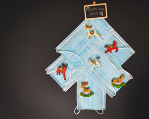 View of a Christmas tree made of blue surgical masks decorated with colorful Christmas vintage wooden ornaments against black chalkboard background. Christmas during pandemic or medical background.