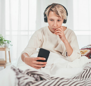 Young woman sitting on cozy bed dressed pajamas browsing the internet and listening music using wireless headphones. Music playing modern technology and free time spending concept