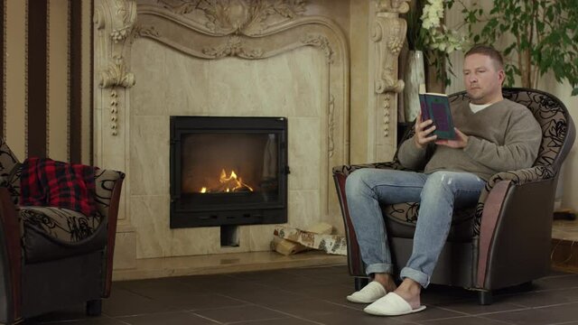 Man reading book near fireplace at home. Video. Middle aged man sitting in front of fireplace in his armchair at home on a cold winter day, reading book.