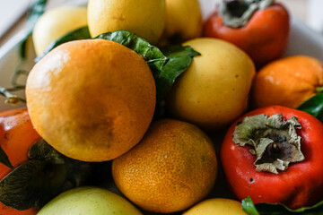 Brightly colored, orange and yellow, winter fruit: persimmon, apples, lemons and oranges arranged on a platter. Some green leaves. 