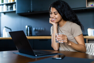 Smiling afro woman with glass of water using laptop in the kitchen at home