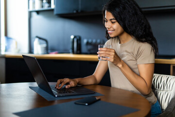 Obraz premium Smiling afro woman with glass of water using laptop in the kitchen at home