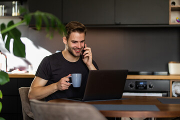 Young handsome man working from home in the kitchen with a laptop talking on mobile and smile