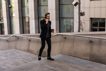 Business talk on a go. Confident young manholding coffee cup and talking on mobile phone while walking outdoors