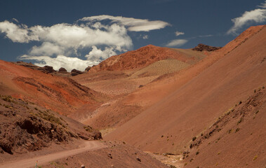 Desert road across the arid Andes mountain range. Traveling along the dirt route very high in the cordillera. The v shaped valley, red and brown mountains, sand and rock under a beautiful sky.