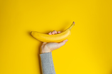Female hand in gray clothes holding one banana on yellow background. Creative banner with two colors of the year 2021 - Illuminating and Ultimate Gray.