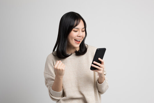 Happy Young Asian Woman Looking At Screen Of Smartphone Standing On Isolated White Background. She Very Happy.