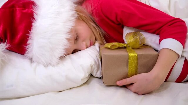 Little girl in Santa hat and Christmas pajamas lying in bed with gift box at home. New Year and Holidays. Christmas morning happiness
