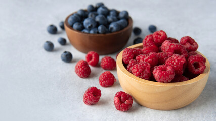 Closeup focus fresh raspberries with blurred blueberries in a wooden bowl soft grey blue background.