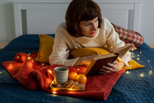 Beautiful mature woman reading book. Cozy everning relaxing and reflection. Stay home concept. Christmas decoration with garland lights, pillows and hot drink with cookies on bed. Dark twilight