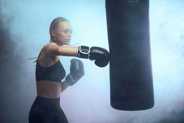 sporty brunette woman in boxing gloves and sportswear kicking bag on dark background with smoke.