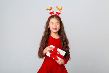 Cute little Asian girl in a red dress holds a gift box on a white background. Christmas concept, text space