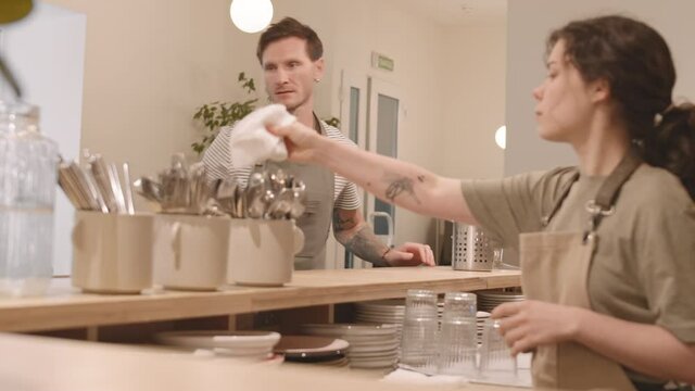 Side view medium close-up of blurred female bartender standing behind counter, polishing forks with white napkin, middle-aged Caucasian man bringing her cutlery stand