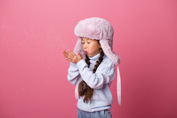 cute little Asian girl in winter clothes blows snow from palms. Winter end, pink background, space for text