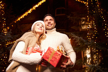 Happy cheerful female and male enjoying great Christmas evening with present boxes in hands in front of their house