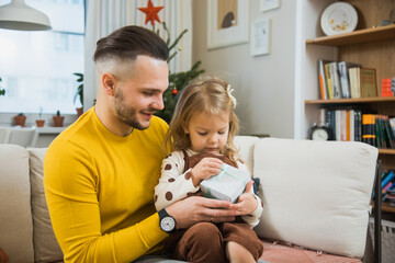 Obraz premium Young father give gift box to little toddler daughter on Christmas. Man and little girl sit on couch together near decorated fir tree.