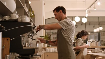 Side view of middle-aged Caucasian man wearing apron using steam wand on massive professional espresso machine in bar, cleaning it, wiping water out, female coworker passing by him and leaving