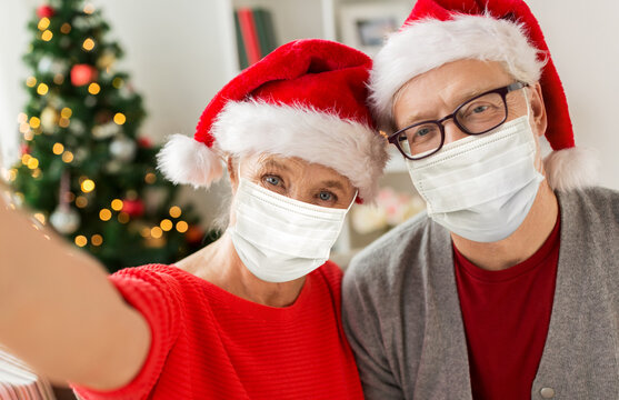 christmas, holidays and health concept - happy smiling senior couple wearing face protective medical mask for protection from virus disease in santa hats taking selfie at home