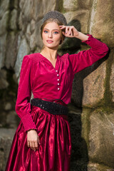 Girl in Red Dress and Silver Tiara Posing with Lifted Hand Near Old Wall Outdoor.