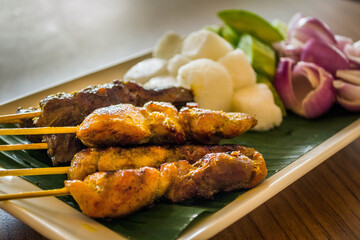 a plate of chicken and beef satay with raw onions, cucumber, rice cake and peanut sauce on the side