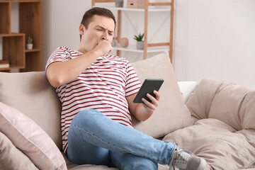 Obraz premium Tired young man yawning while relaxing on sofa and using digital tablet at home, sleep deprivation and leisure time