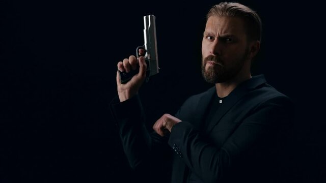 Aggressive bearded man in black clothing using gun while standing in studio. Isolated over black background, nasty look.
