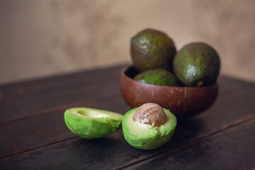 Whole avocado fruits in a bowl made from coconut shells and cut in half lie next to it on a dark wooden background