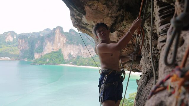 4K Confidence Asian man climber preparing for climbing up the rocky mountain with safety rope and harness at tropical island. Healthy male enjoy active lifestyle and extreme sport in holiday vacation.