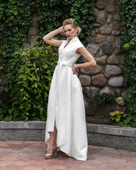 An elegant blonde model in a long white wedding dress and beige shoes poses against a stone wall with green plants. Exterior wedding photography on a summer day