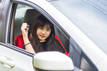 Close up portrait smiling young Asian woman is holding a car key, she is sitting in a white car and looking at the camera