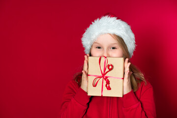 pretty young school girl with Santa Claus hat and red sweater is holding a gift package in front of a red background