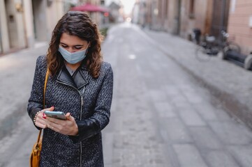 Close-up caucasian smiling young woman walking in a city center using smartphone during cold winter season. Lady wearing a protective face mask. Pandemic, new normal concept.