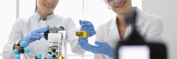 Two women in uniform and chemical goggles hold glass flask of yellow liquid in front of camera. Videography chemical experiments blogging concept.