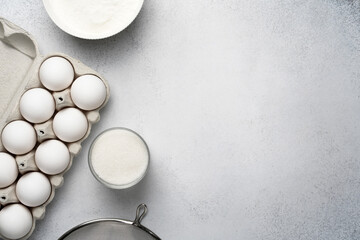 Wheat flour, sugar, sieve and white eggs on the grey background. Baking ingredients for cake. Top view, copy space.