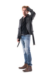 Confident young stylish man in leather jacket and jeans touching hair style looks at camera. Full...