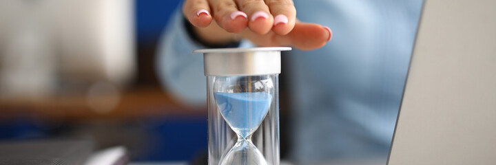 Woman holds her hand over hourglass at workplace in office closeup. Time is money concept.