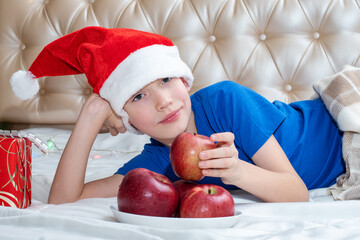 Fototapeta na wymiar Healthy food concept for Christmas. Cheerful cute Caucasian boy in Santa hat lies on the bed next to a plate of apples and a gift. Boy takes an apple as a symbol of healthy eating