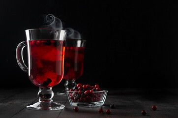 Christmas bright red mulled wine with berries, steam, ripe cowberry, blueberry in two glass wine glasses on black wood table in dark interior, copy space..