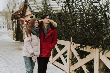 Beautiful young caucasian heterosexual couple of shoppers are choosing and making a purchase at the Christmas tree market in winter outdoors in snowy weather