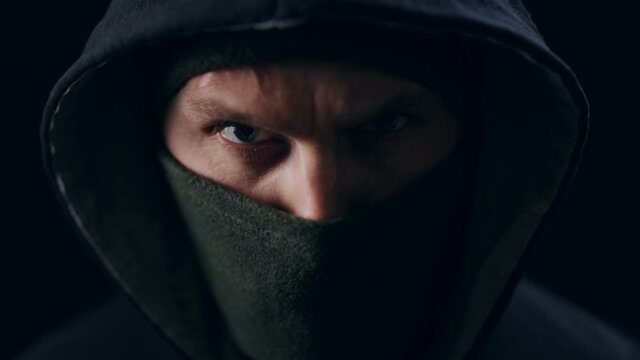 Close up of bad guy wearing black mask and hood while looking with penetrating glance. Isolated over black studio background. Concept of crime.